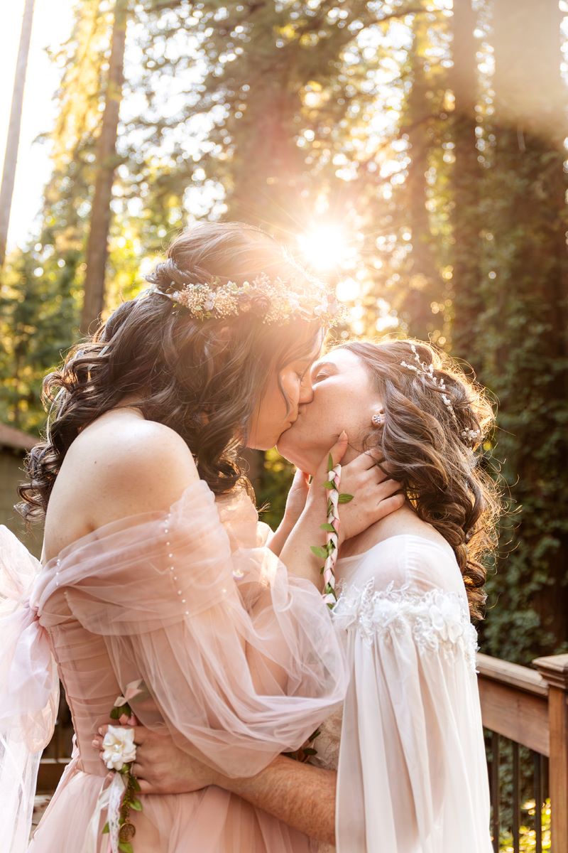 A couple kissing on a wooden deck surrounded by the forest one is wearing a pink gown and flower crown the other is wearing a white gown 