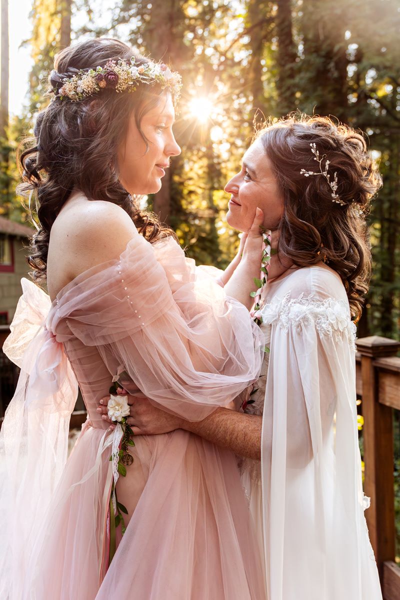 A couple looking into each other's eyes on a wooden deck and smiling at each other one is wearing a white gown and the other is wearing a pink gown with a flower crown 