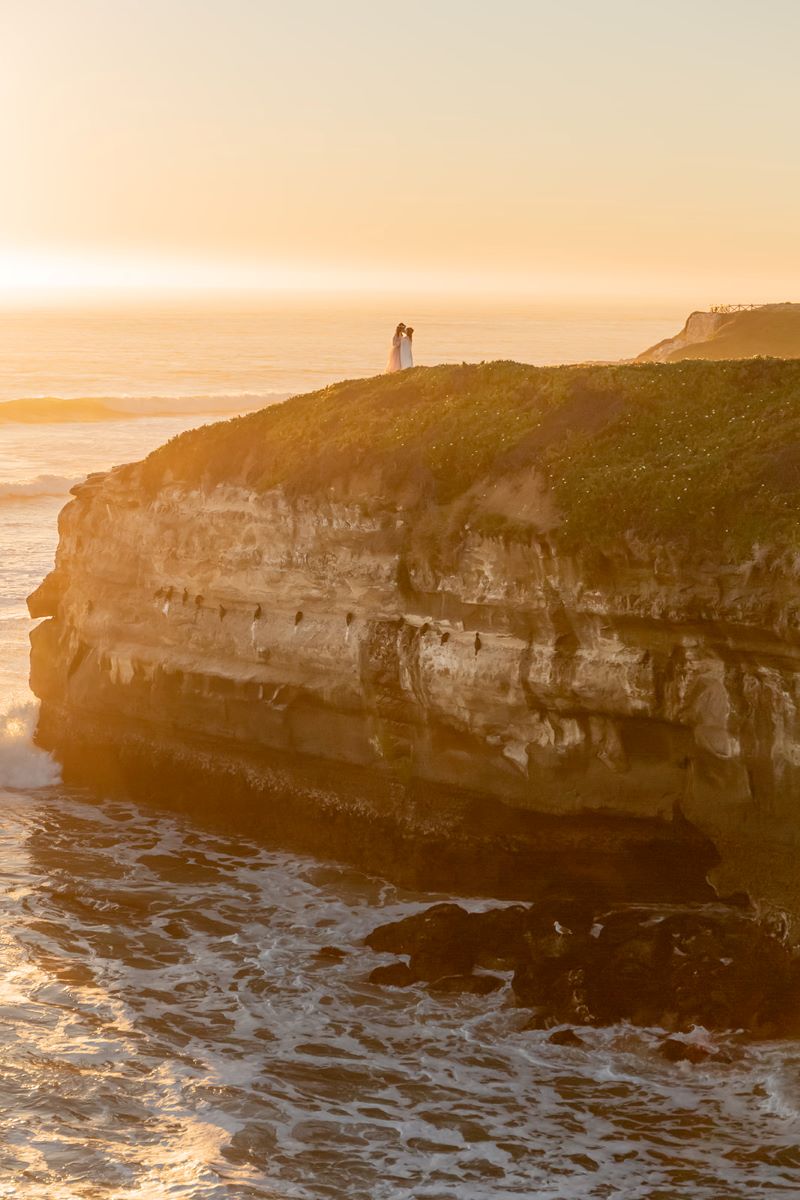 A couple kissing on a cliffside at sunset next to the ocean one is waring a pink gown the other is wearing a white gown 