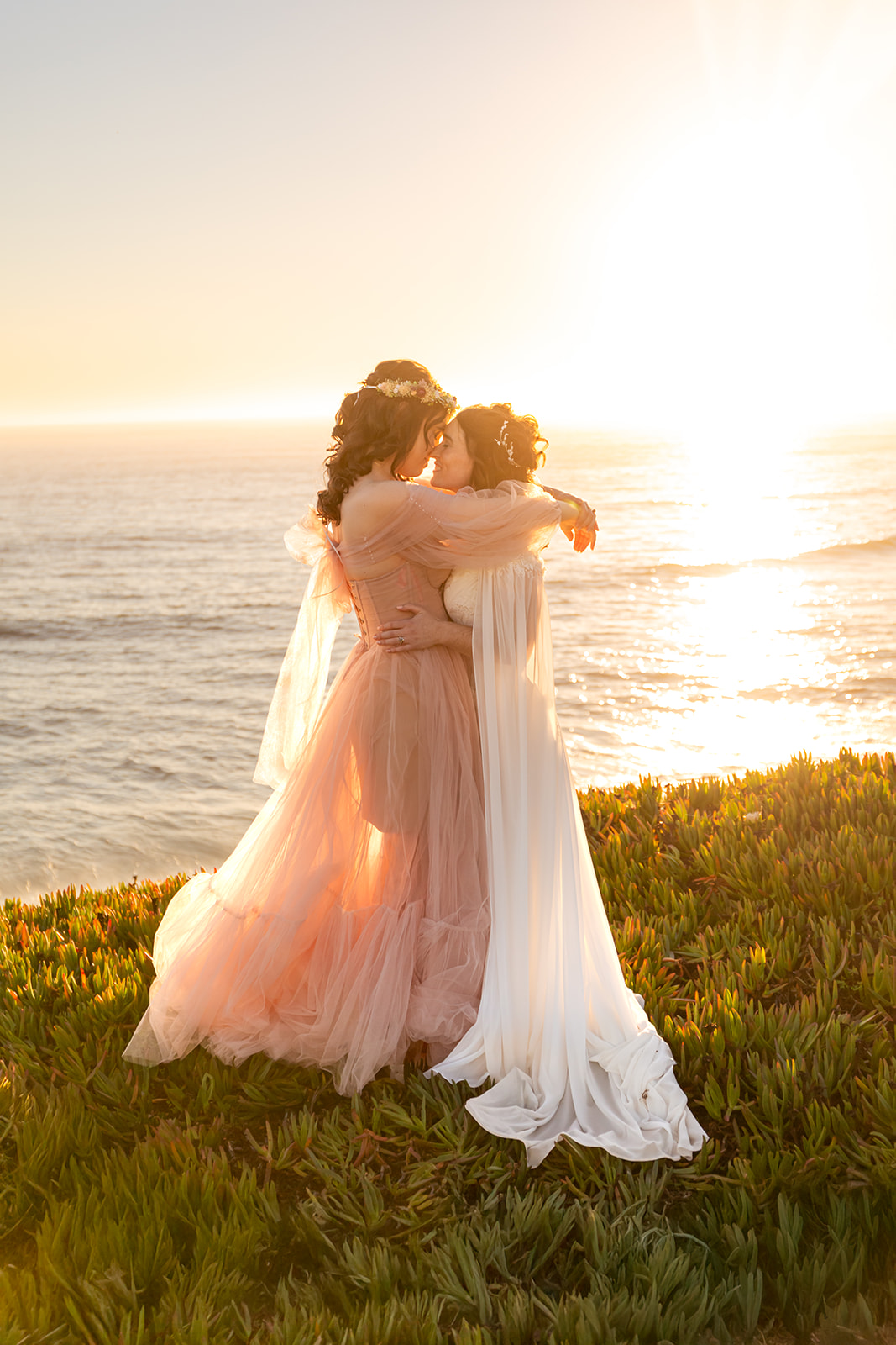 A couple pressing their foreheads to each other on a cliffside at sunset next to the ocean one is wearing a pink gown and has their arms draped on their partner's shoulders the other is wearing a white gown and has their hands on their partner's waist 