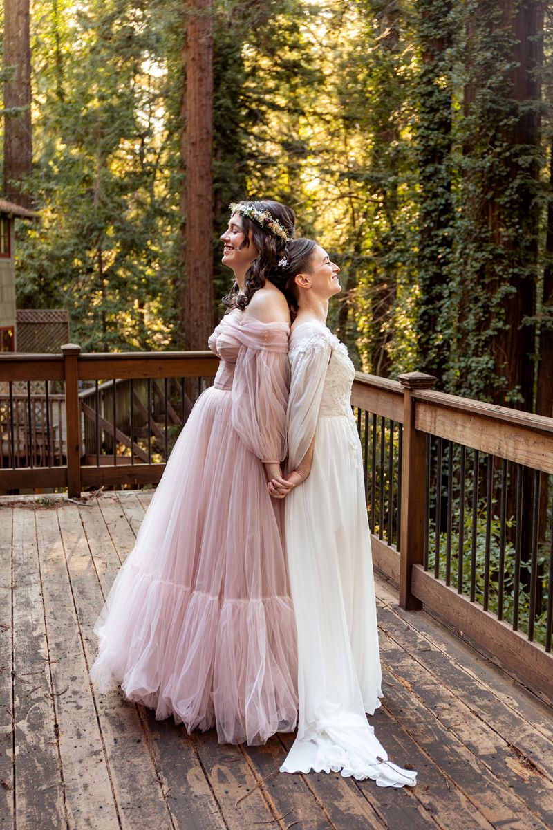 A couple standing on a wooden deck with their backs to each other and holding hands one is wearing a pink gown and the other is wearing a white gown