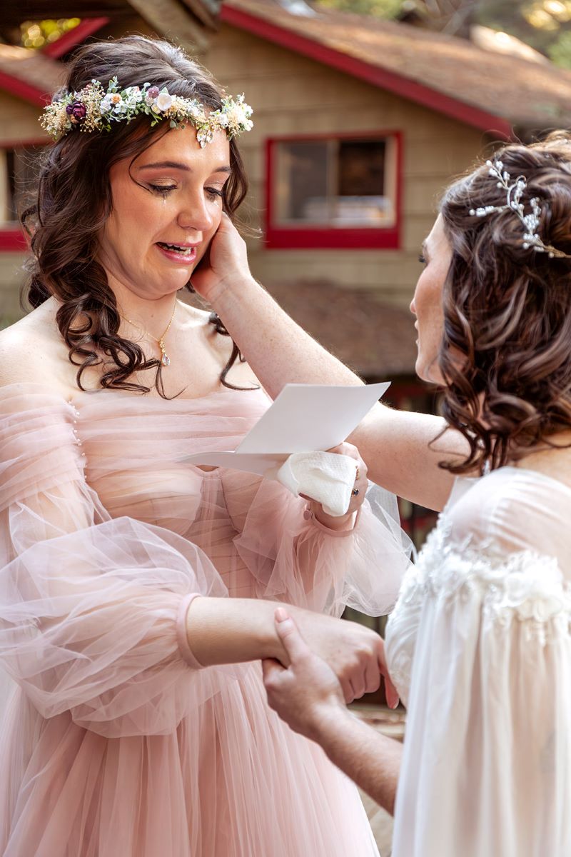 A couple at their elopement ceremony one is wearing a pink gown and reading their vows to their partner the other is wearing a white gown and holding their partner's hand with one hand and touching their face with the other 