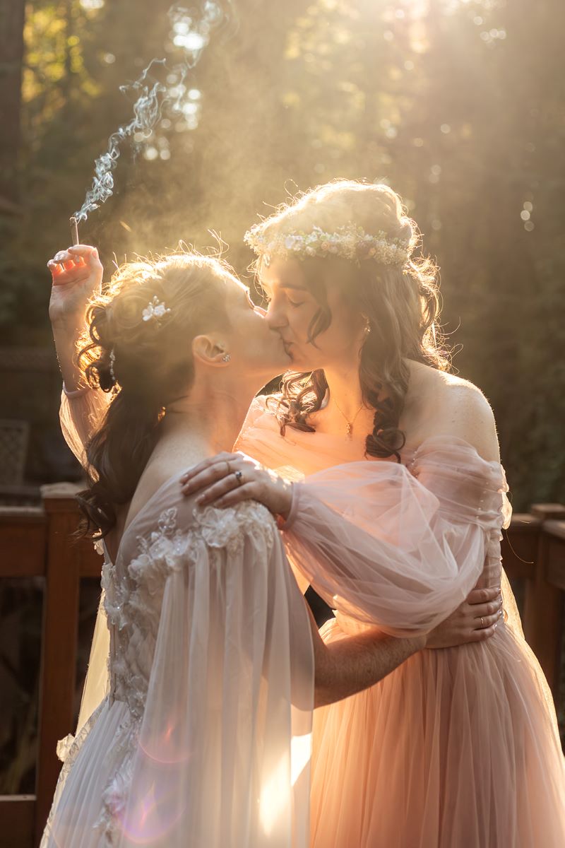 A couple kissing on a wooden porch deck one partner is wearing a pink gown and holding a joint the other is wearing a white gown and touching their partner's waist 