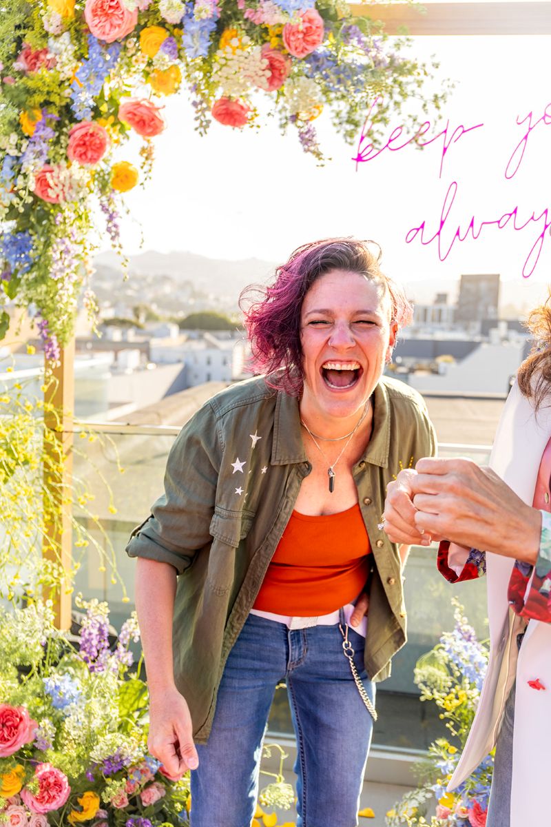 a couple standing in front of a beautiful floral arch one is looking towards the camera laughing and smiling with excitement