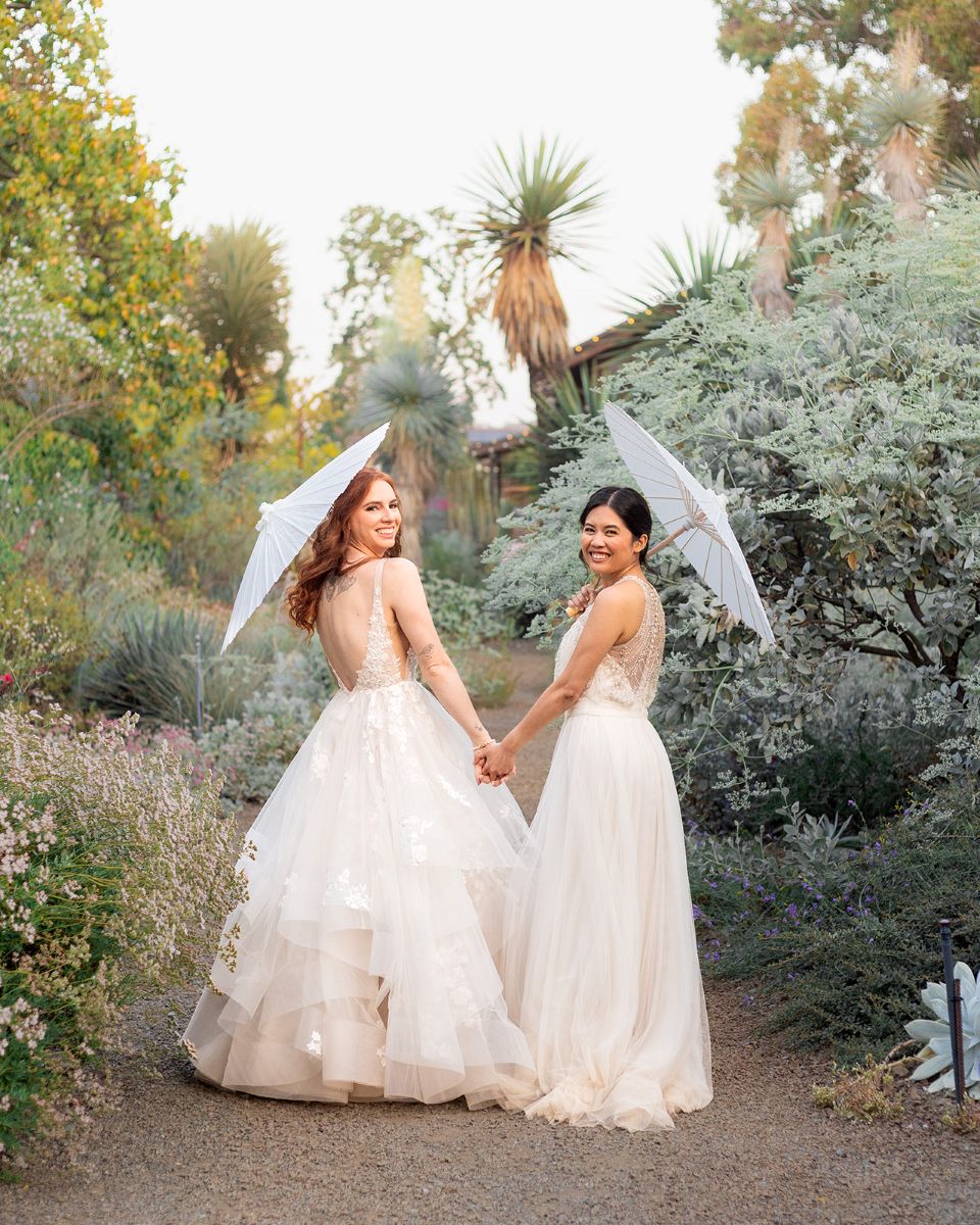 two women holding hands and white parasols and wearing white wedding dresses standing in a garden