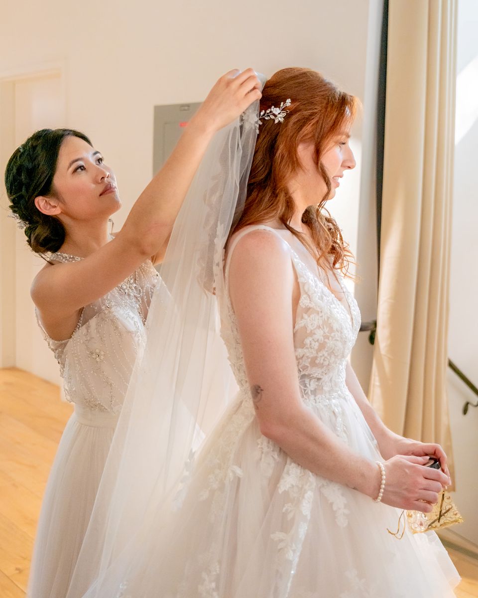 two women in white wedding dresses one is placing a veil on the other's head