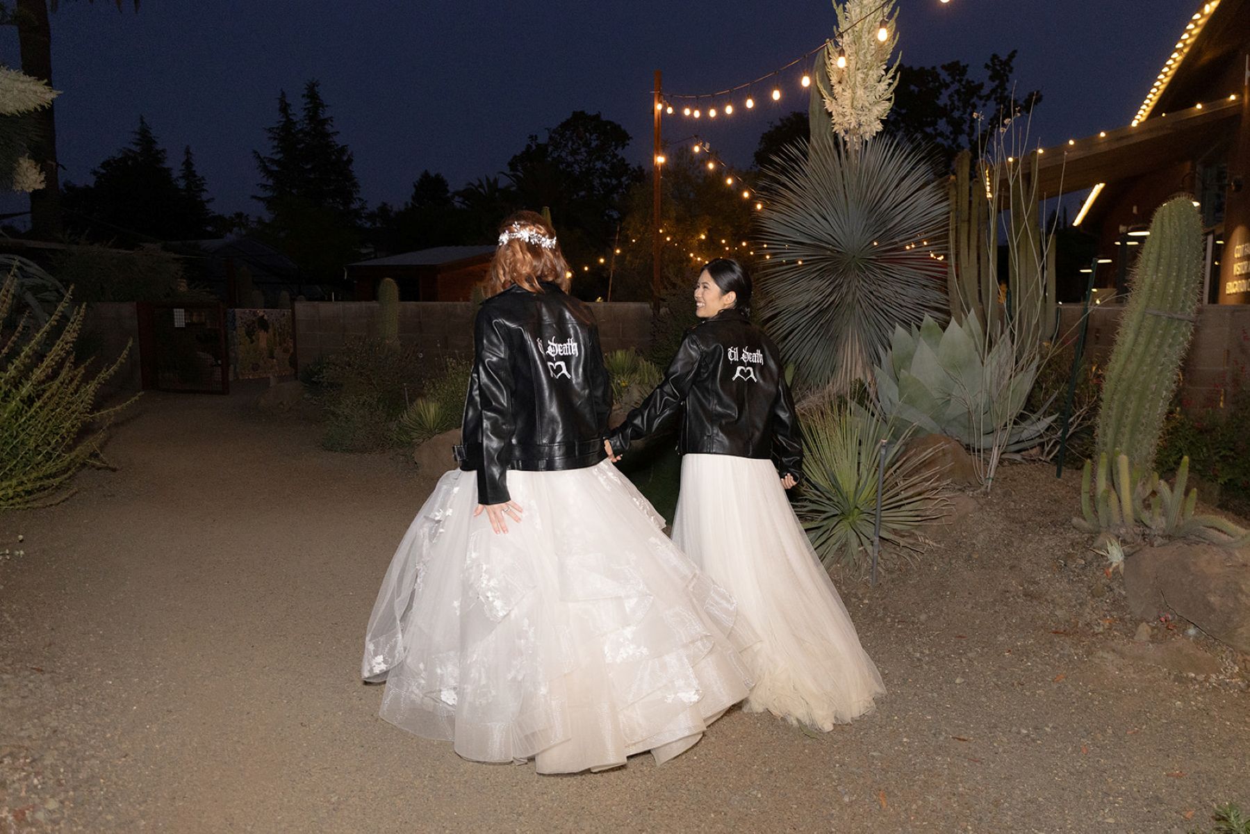two women wearing white wedding dresses and black leather jackets walking through the ruth bancroft garden at night