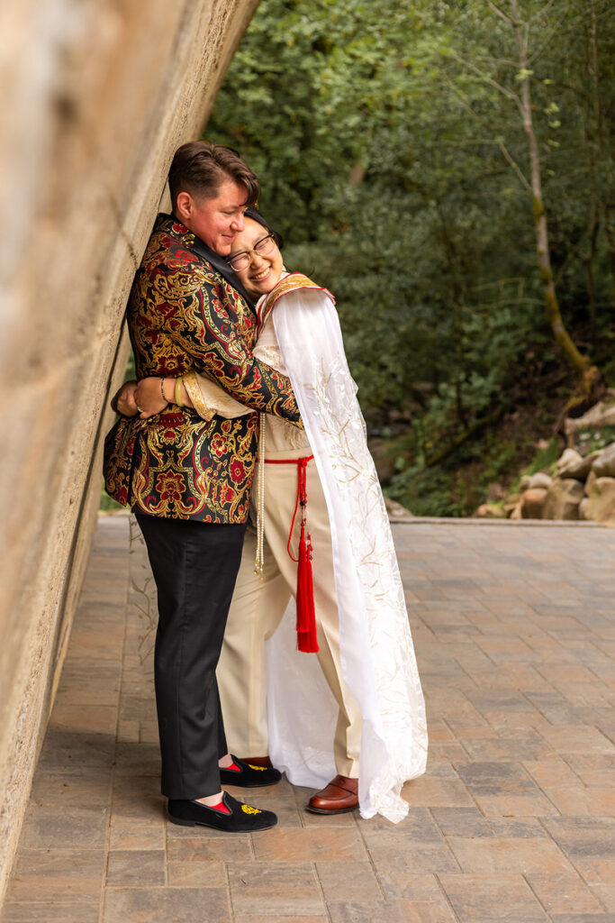 A newlywed couple hugging and leaning against an arched bridge