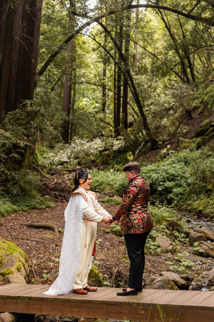 A newlywed couple holding hands and standing on a small bridge in the woods