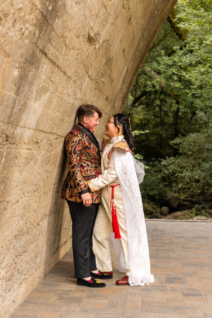 A couple holding hands and smiling at each other as one leans against an arched wall