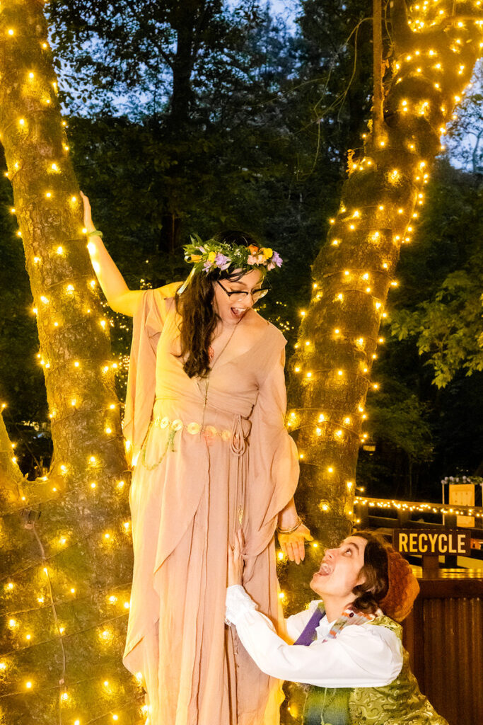 A person hanging on to a fairy light covered tree with another person reaching up to them from below
