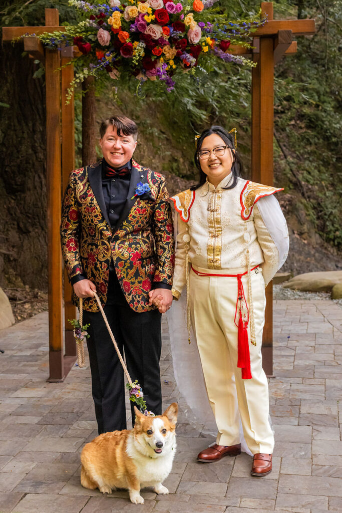 A newlywed couple holding hands and smiling with their dog sitting at their feet