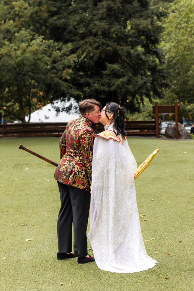 A newlywed couple kissing while one holds an axe and the other holds a sword