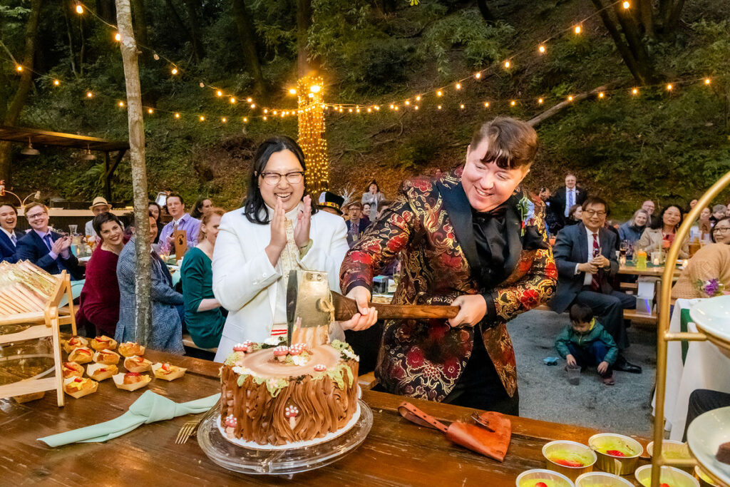 A newlywed cutting a wedding cake with an axe with their partner cheering beside them