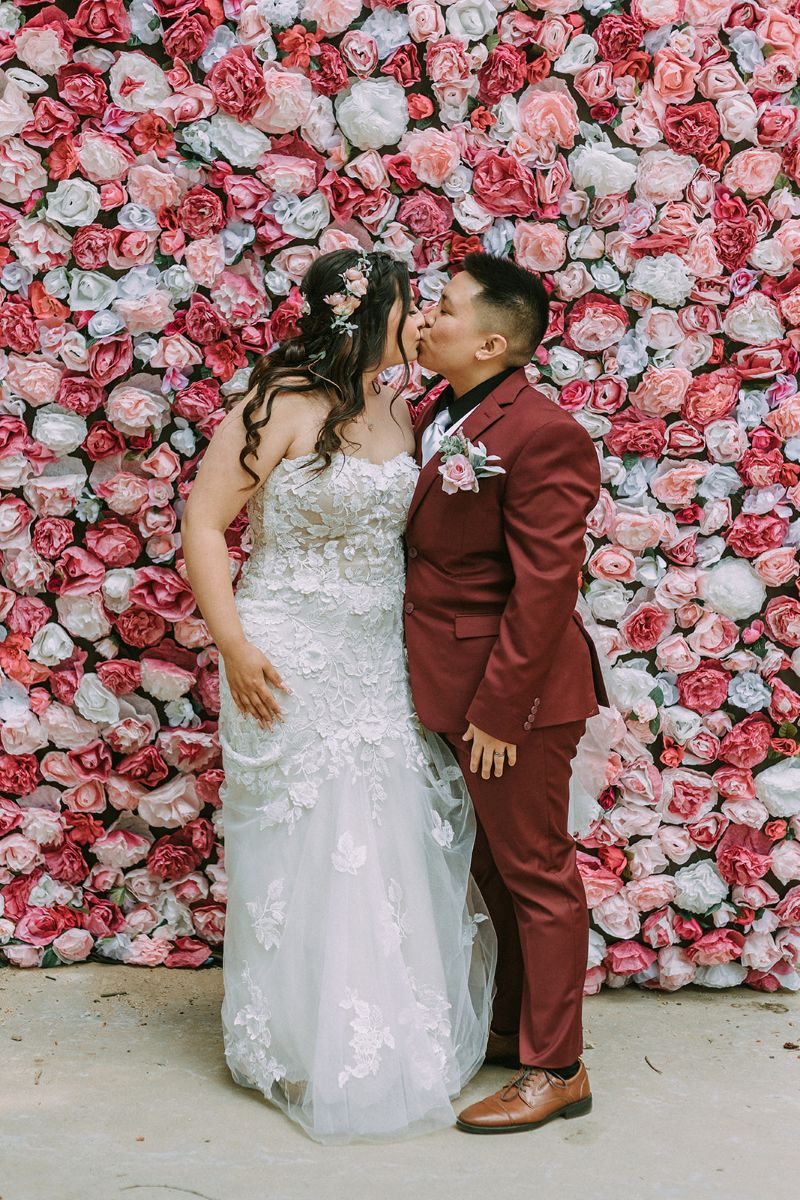 a couple kissing in front of a colorful floral wall made up of dyed coffee filters to make pink and white flowers on the day of their wedding