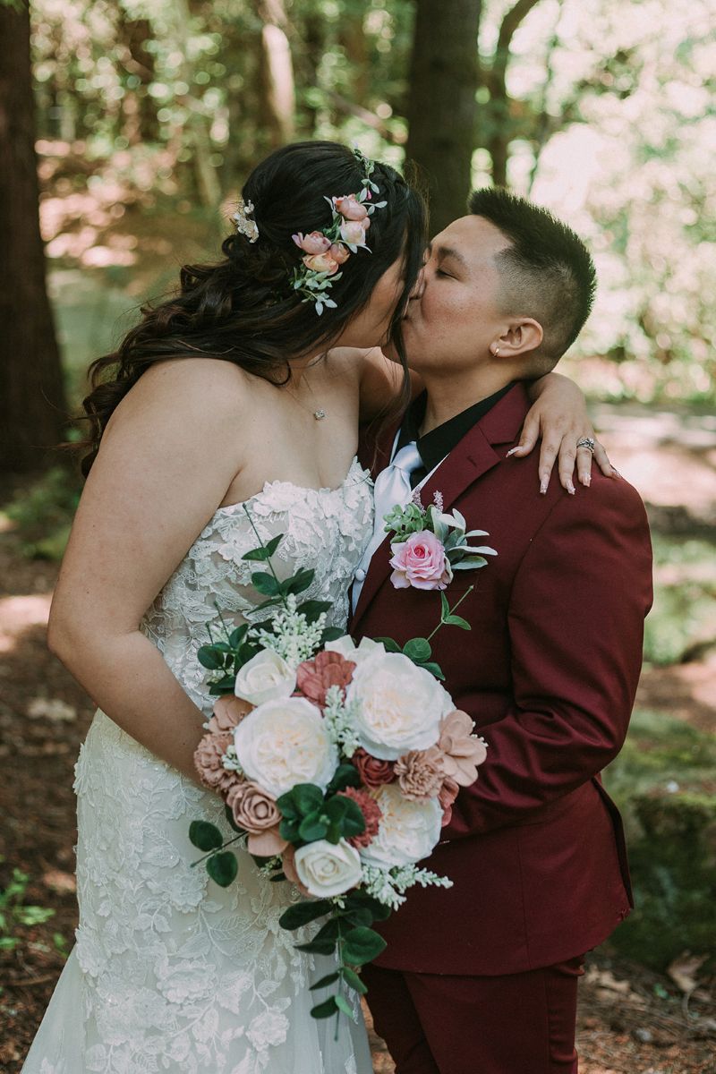 a queer couple kissing each other surrounded by redwoods in saratoga california on the day of their wedding one is wearing a white wedding dress the other is wearing a red suit