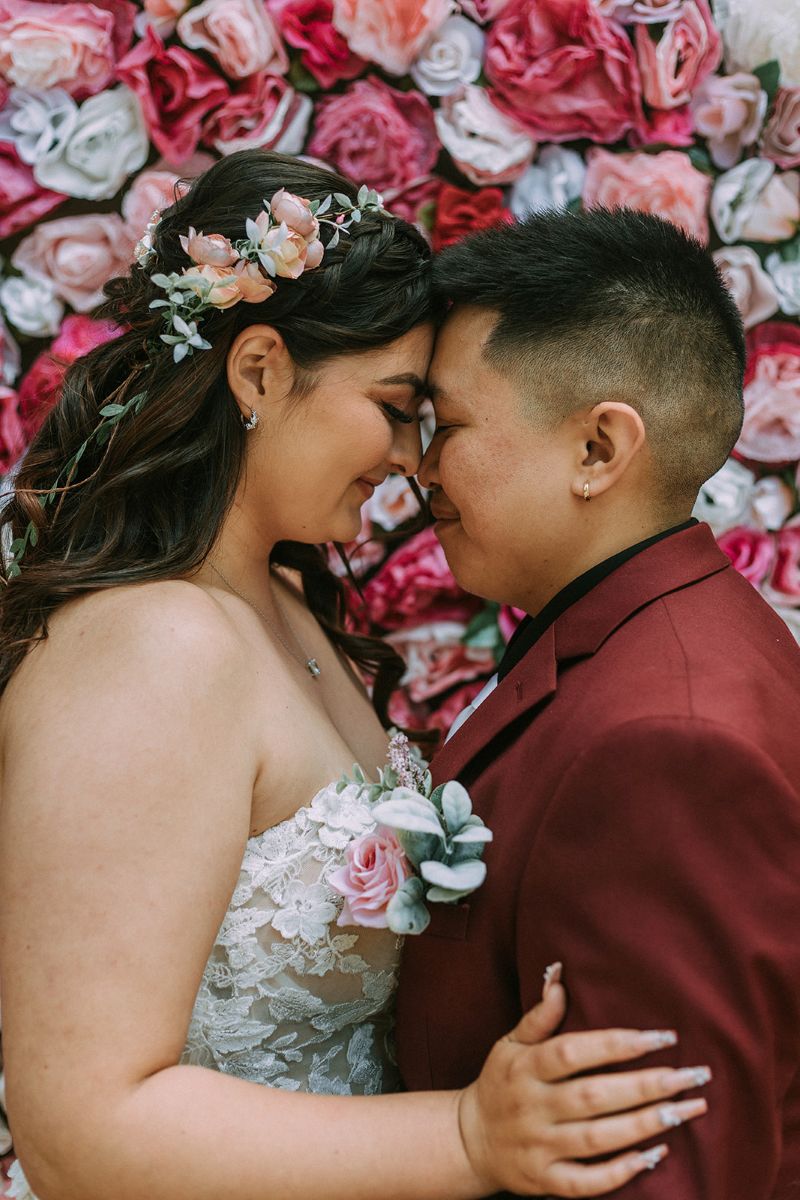 a couple standing in front of a wall of paper flowers with their heads pressed together and smiling at each other on the day of their wedding