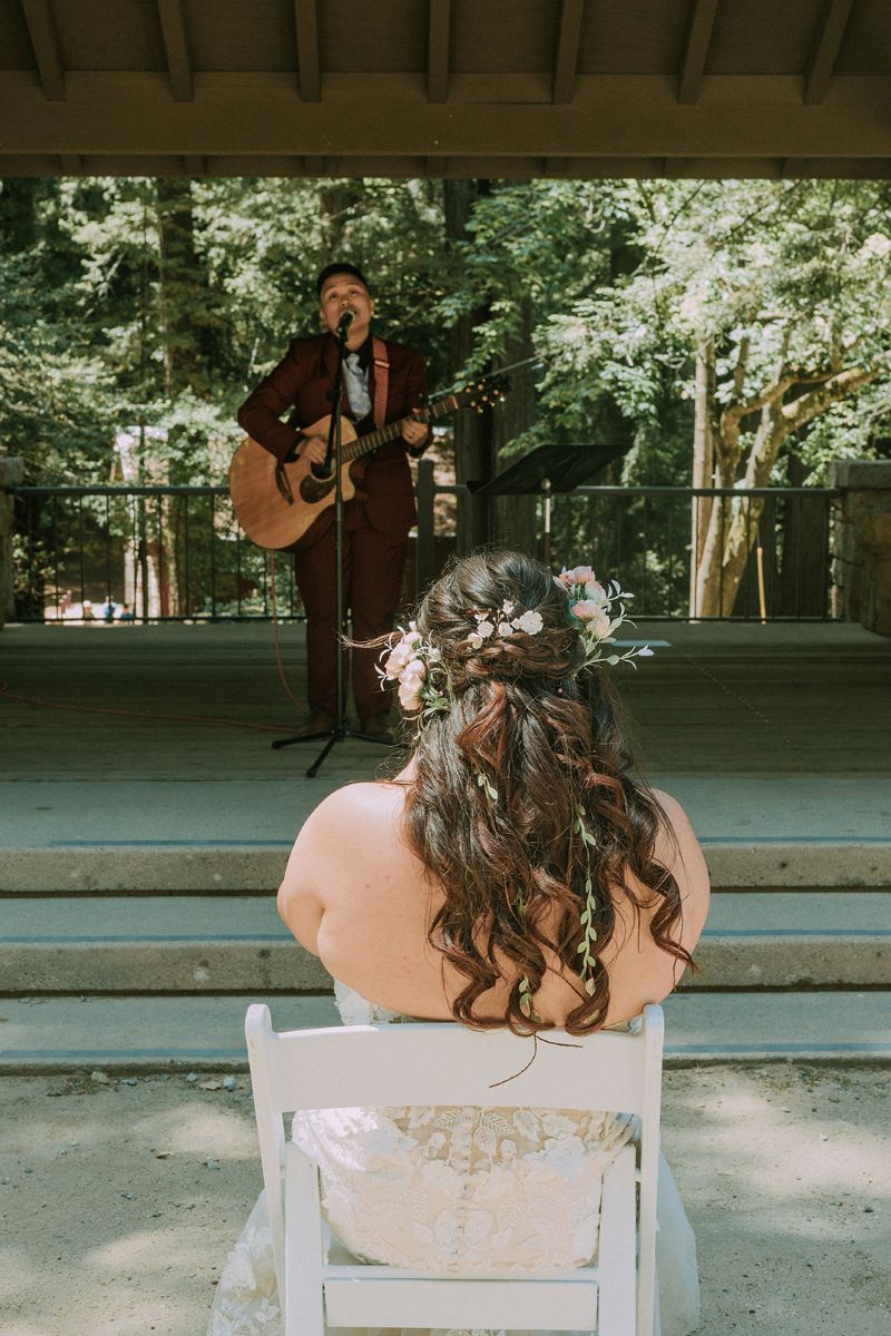 a couple at their wedding reception outdoors one is serenading the other with a guitar the other is sitting in a white chair and watching their partner