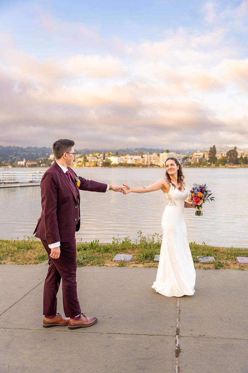 a couple by the water in oakland with the city skyline behind them they are holding hands and looking at one another 