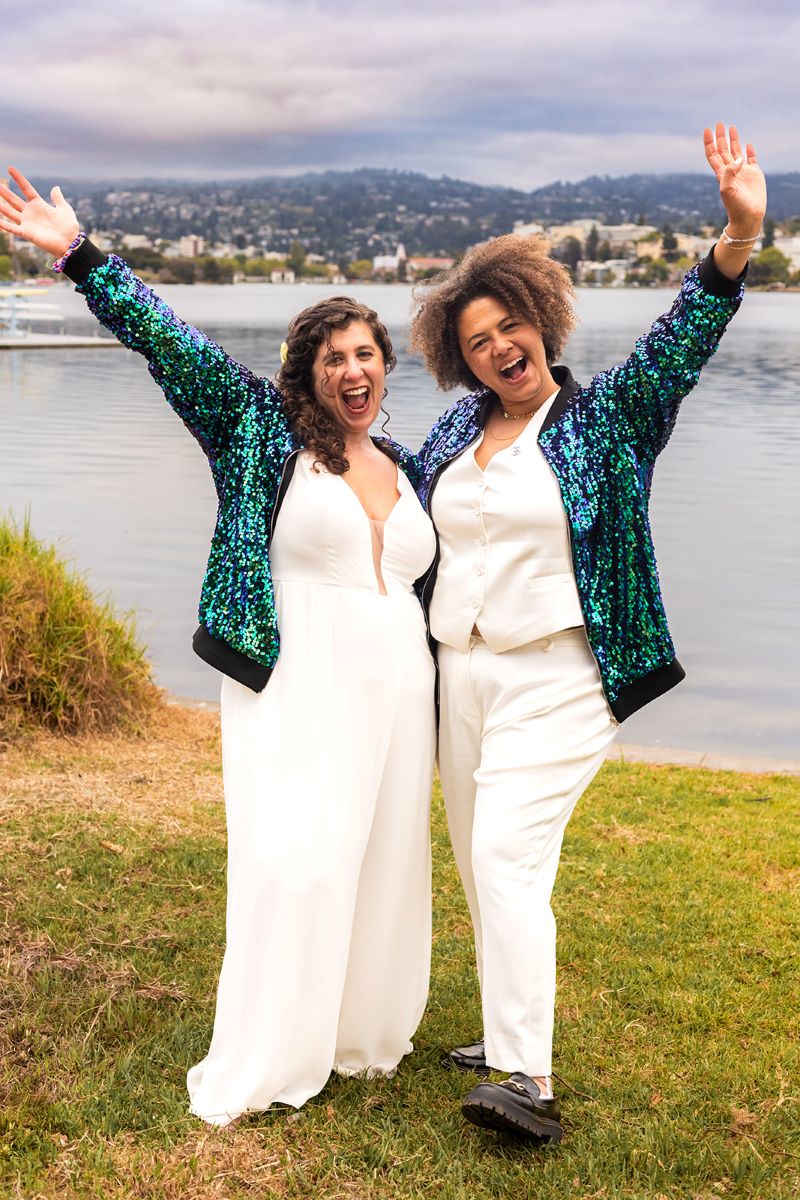 a couple standing next to the water in oakland california wearing matching jackets and they both have their hands in the air and are smiling 