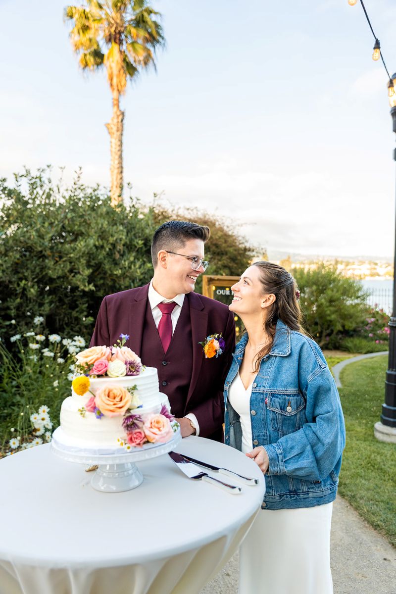 a couple smiling at each other and standing next to their wedding cake at their wedding reception 