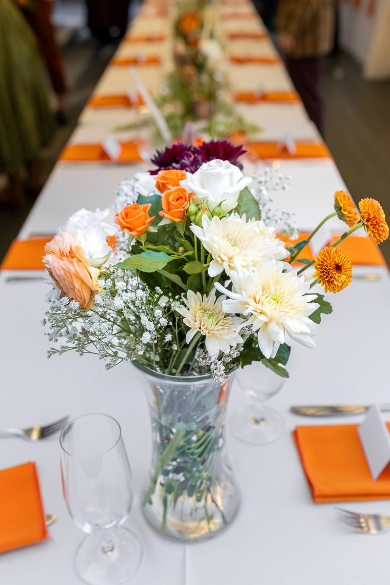 fa mix of orange, white, and violet flowers in a vase on a banquet table set up for a wedding reception the table has a long white cloth on top of it 