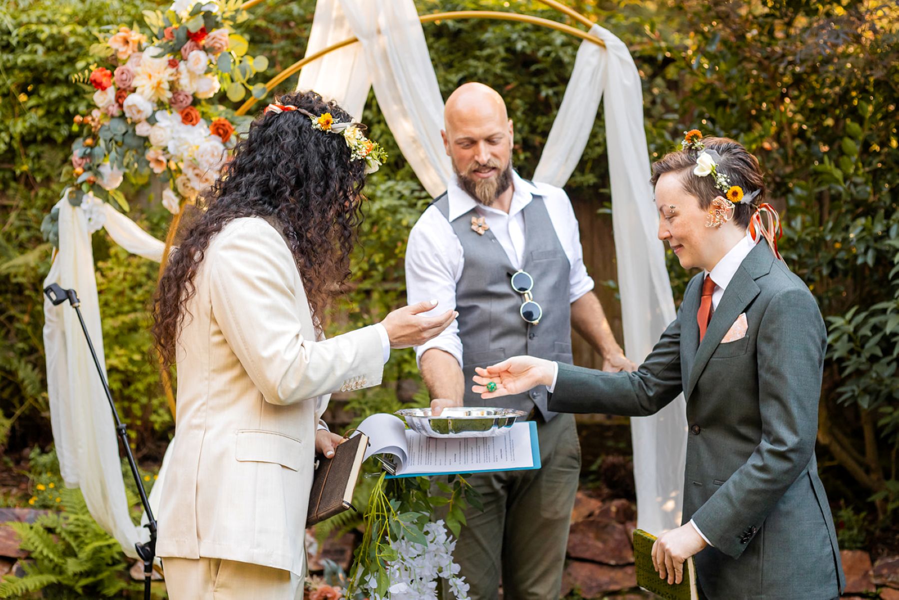 a couple putting dice into a metal bowl at their wedding ceremony their officiant is holding the metal bowl behind them is a moon shaped arch 