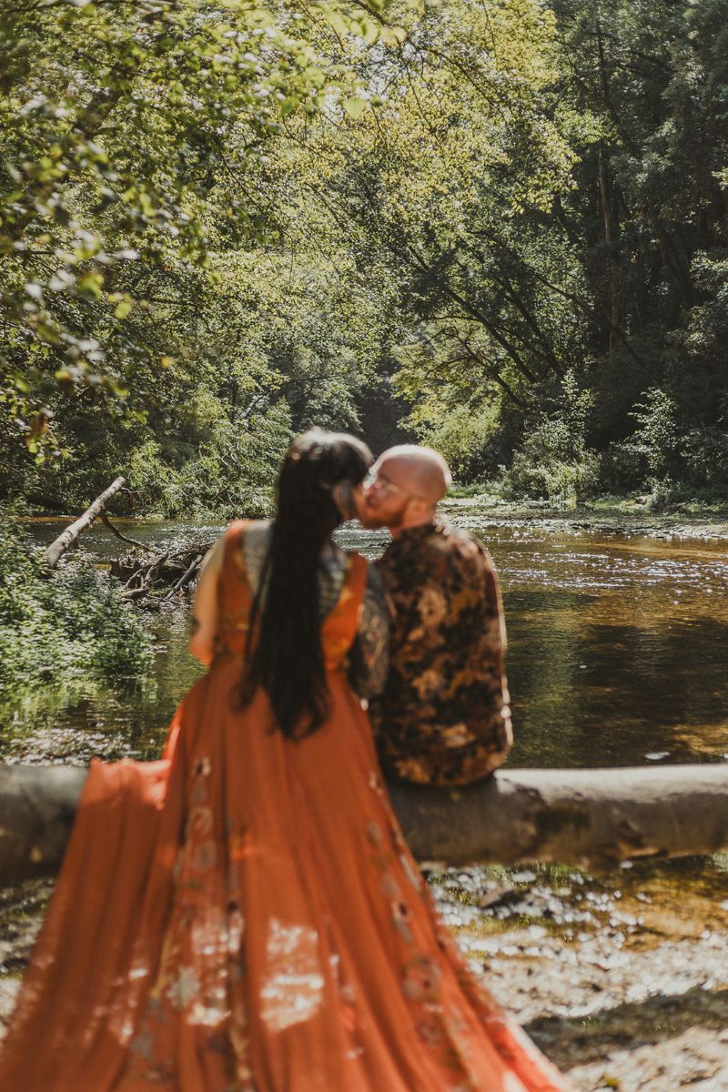 a couple sitting on a log next to the water in henry cowell state park kissing each other 