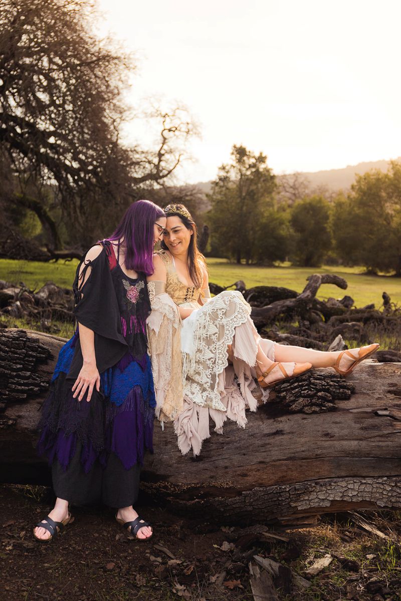 an lgbtq couple wearing fantasy inspired outfits for their wedding and sitting on a fallen tree 