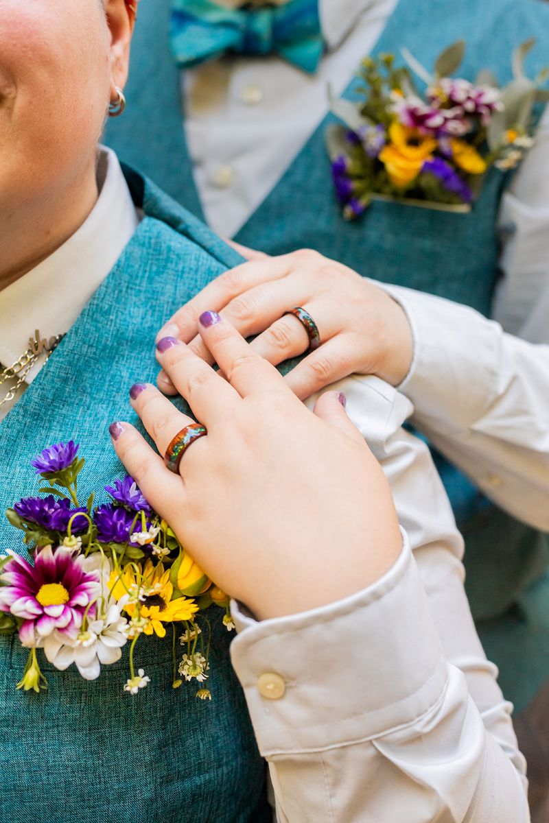 a couple holding hands and showing off their matching rings they are both also wearing matching blue vests and flower pocket inserts 
