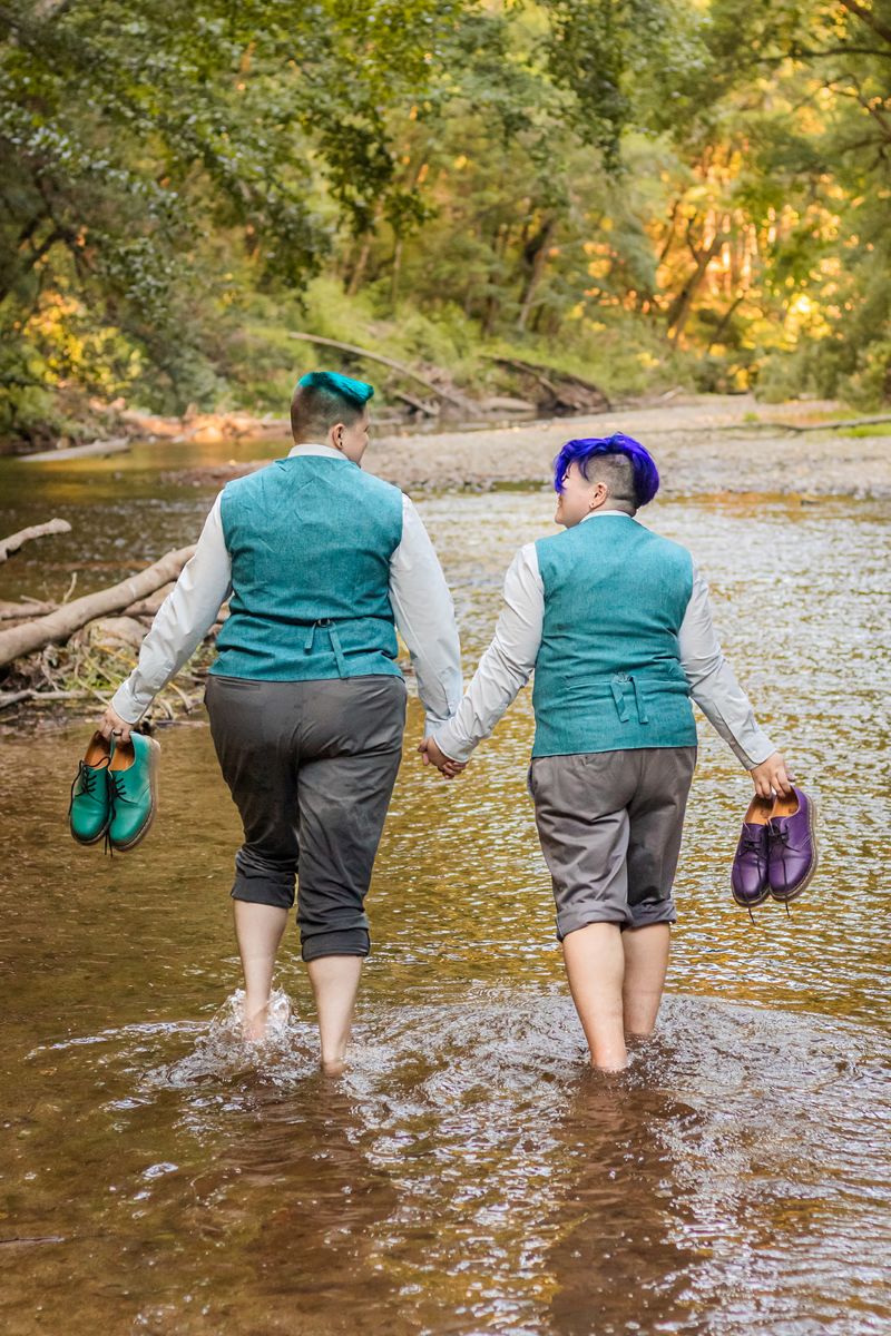 a couple wearing matching blue vests holdind hands and their shoes and walking through a stream together 