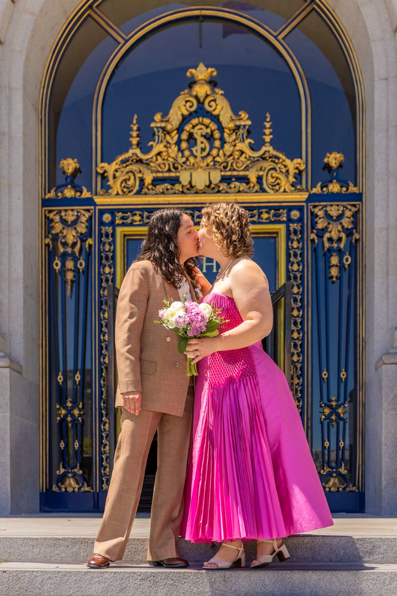 a couple kissing in front of a blue door one is wearing a pink dress and holding flowers the other is wearing a brown suit 