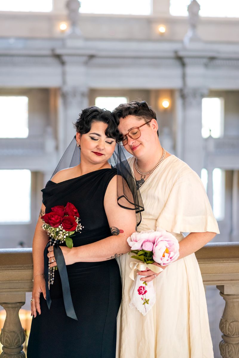 a couple leaning their heads against each other one is wearing a white linen dress and holding pink peonies the other is wearing a black dress and veil and holding roses 
