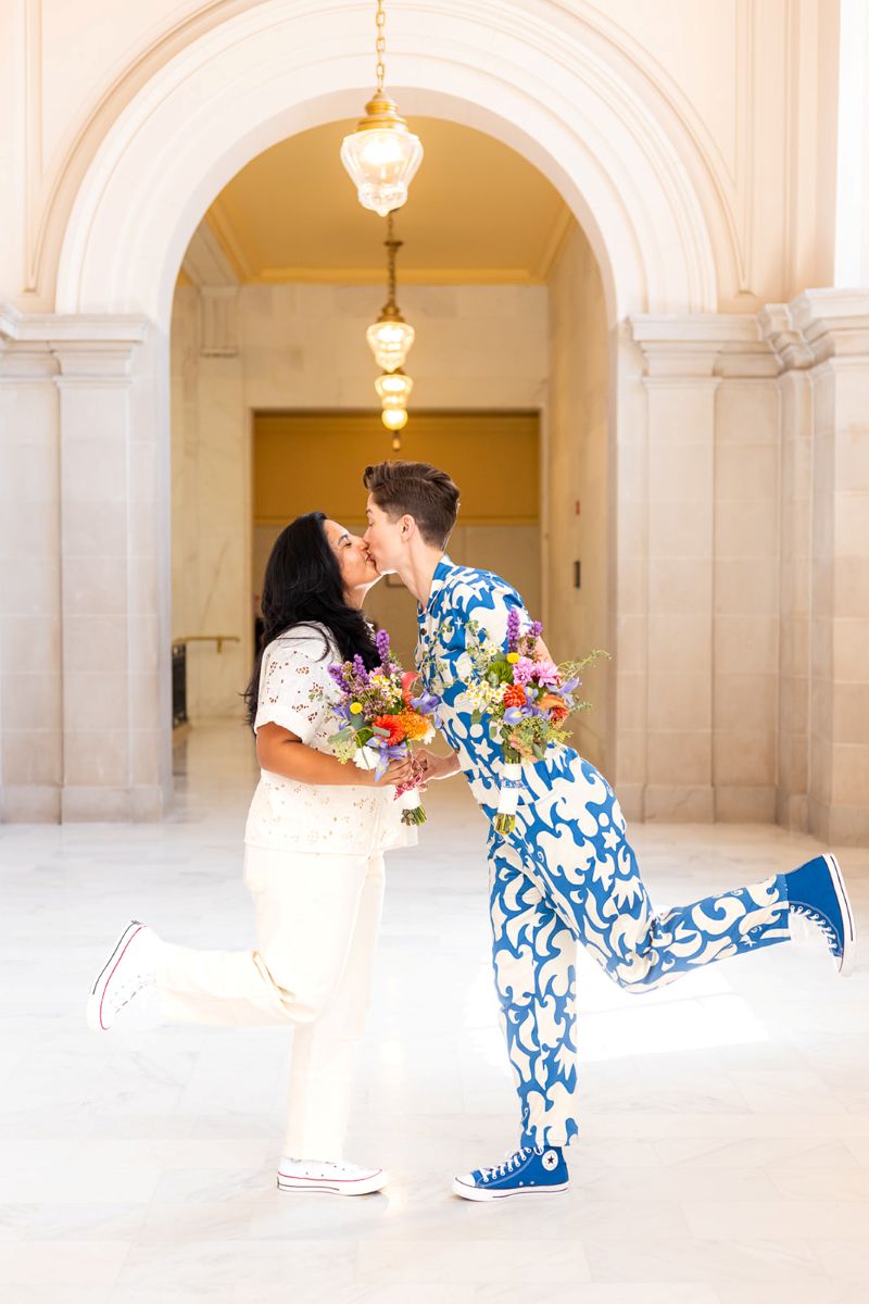 a couple kissing each other in a city hall one is wearing a blue and white jumpsuit with blue converse the other is wearing a white eyelet shirt and white pants and white converse they are also both holding flowers 