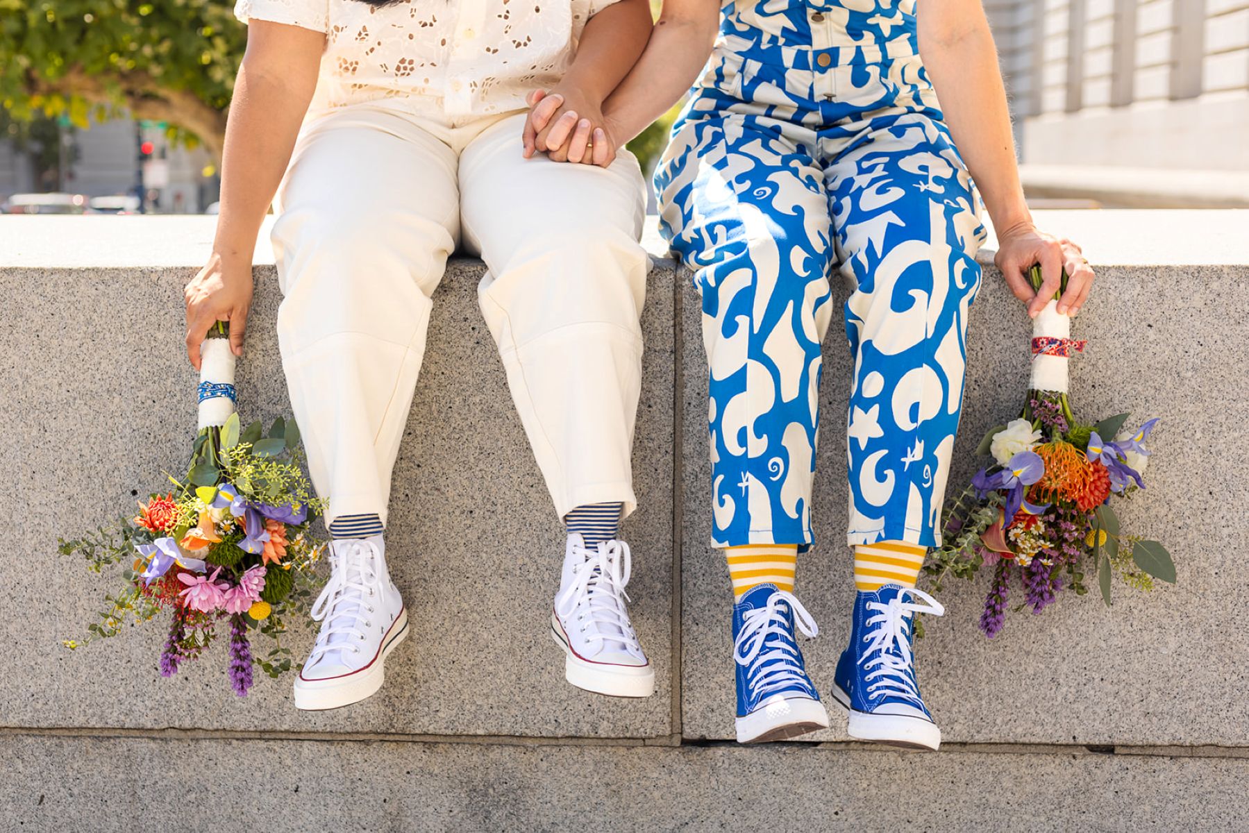 a couple sitting on a concrete wall both holding bouquets one person is wearing blue converse and blue and white pants the other is wearing white pants and white converse 