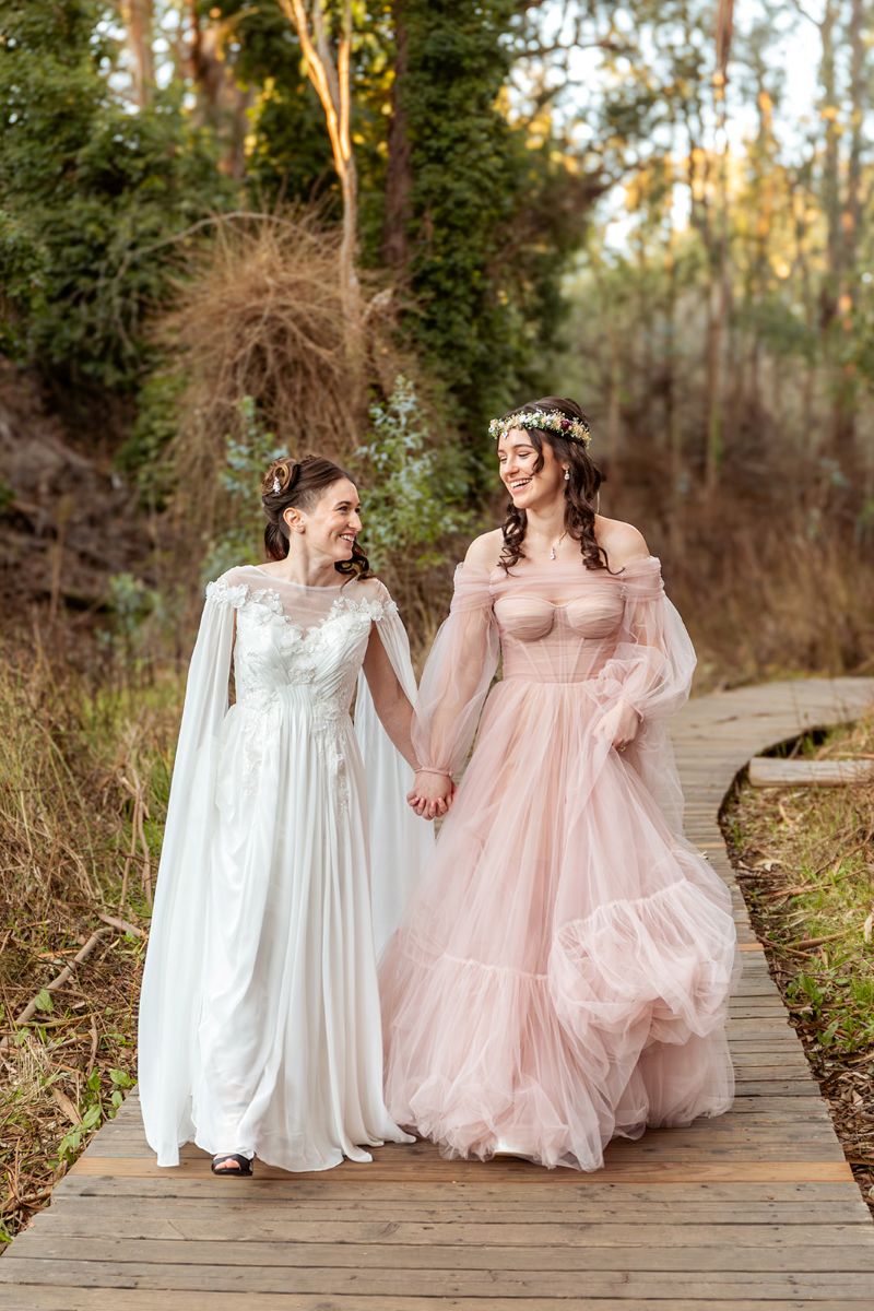 a couple on their wedding day wearing fantasy inspired dresses and holding hands walking on a wooden path 