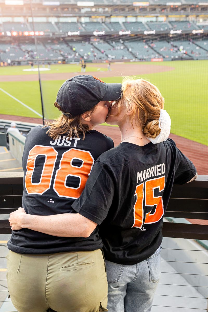 a couple at a baseball stadium wearing matching just married jerseys and kissing 