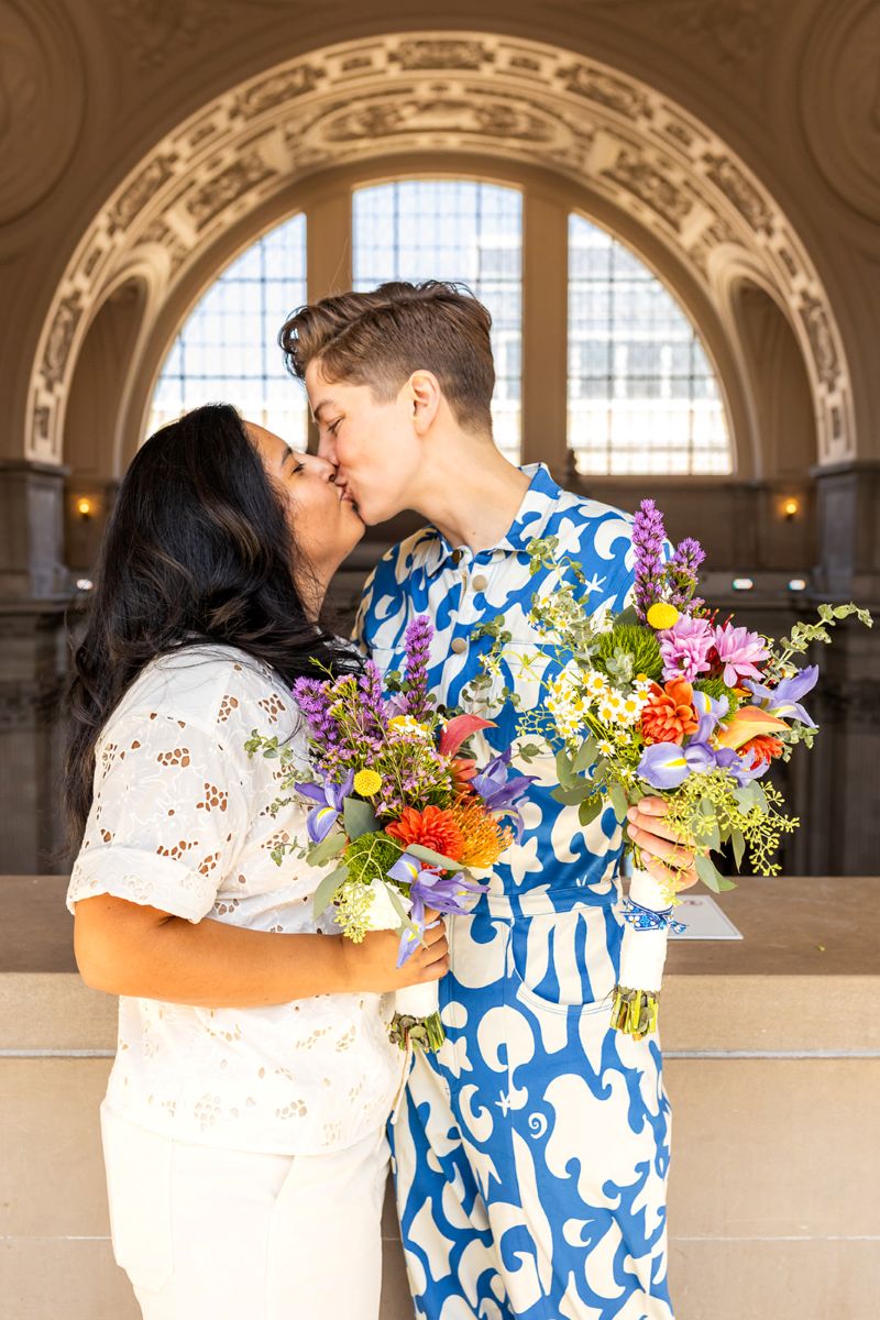 a couple kissing and holding colorful bouquets one is wearing a blue and white jumper the other is wearing a white eyelet shirt 