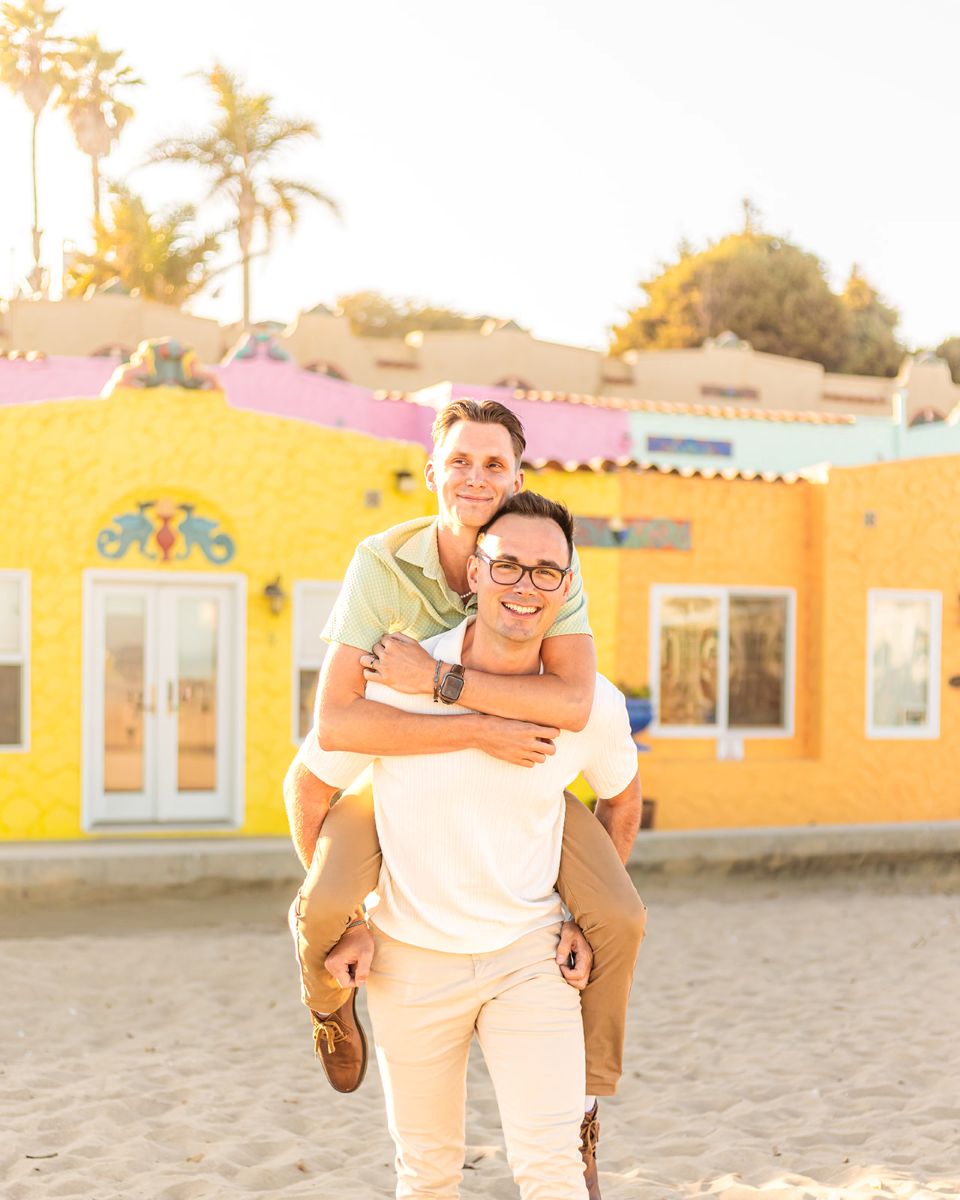 a man has his partner on his back and they are both smiling taking pictures in the sand at capitola beach 