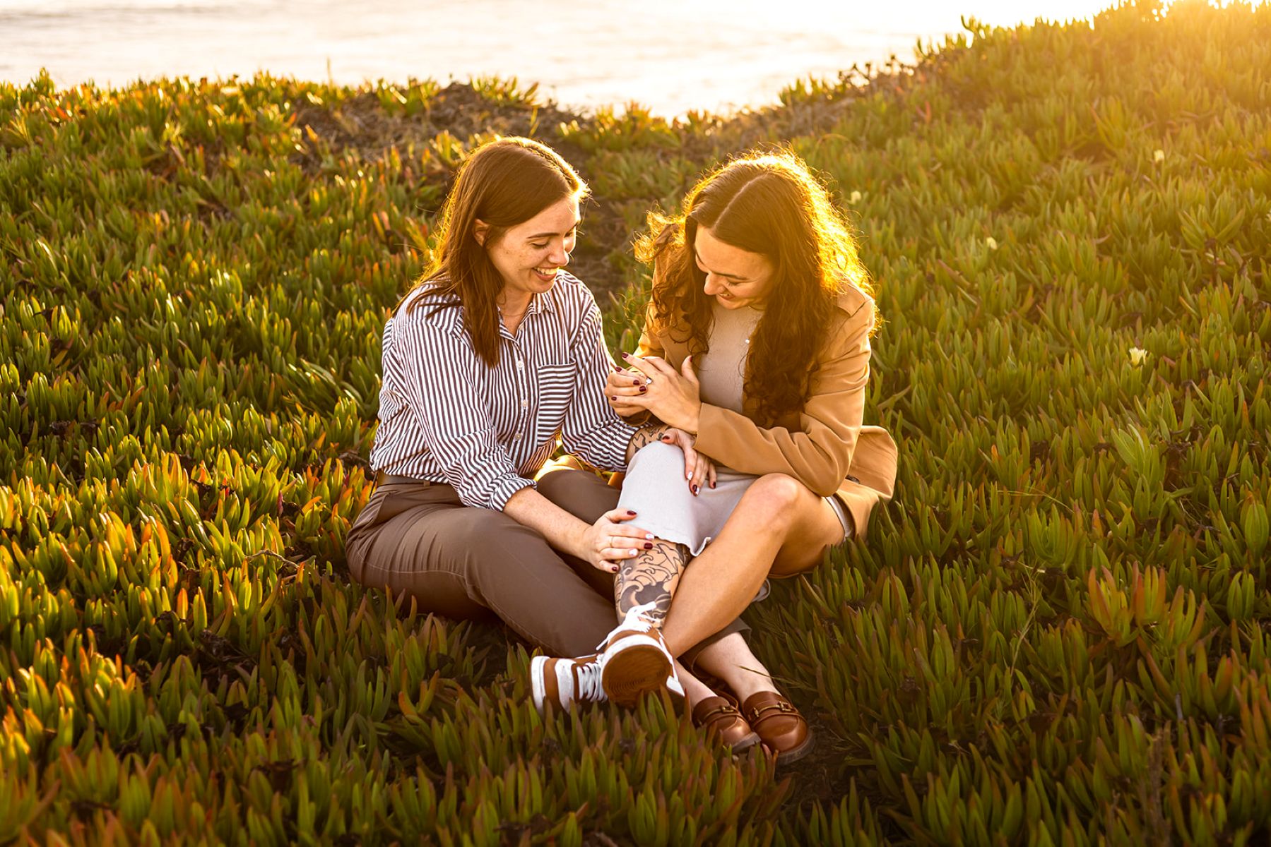 a couple sitting in the grass on west cliff drive next to the ocean at sunset laughing together