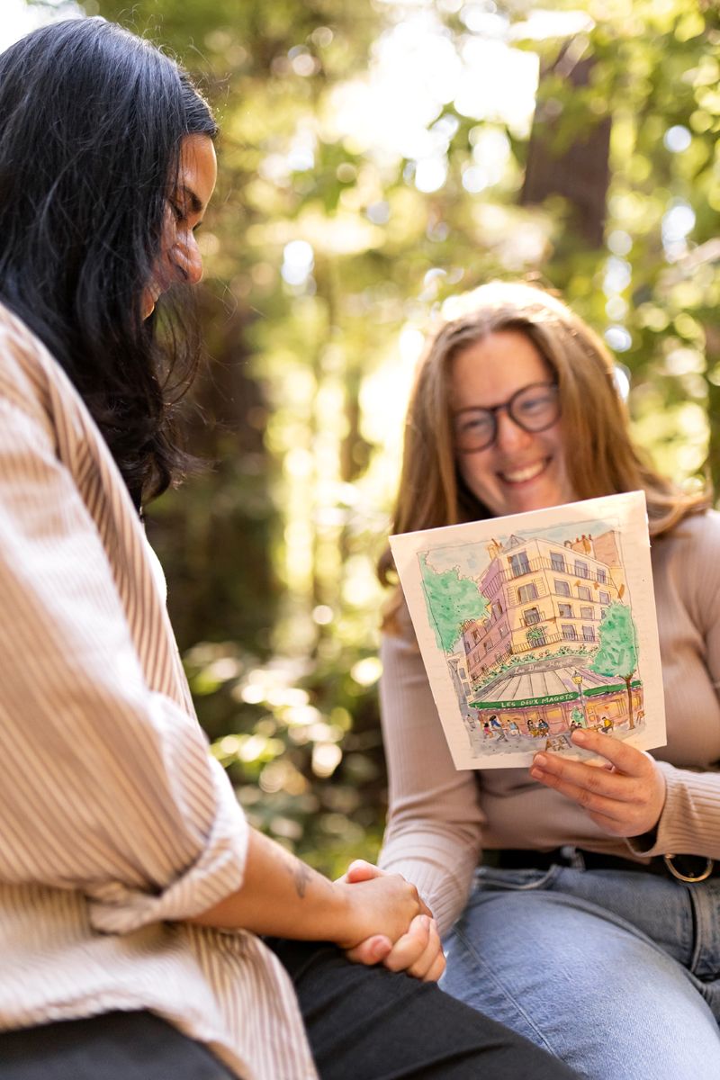 a couple holding hands and sitting on a log in henry cowell state park one partner has gifted the other a watercolor drawing 