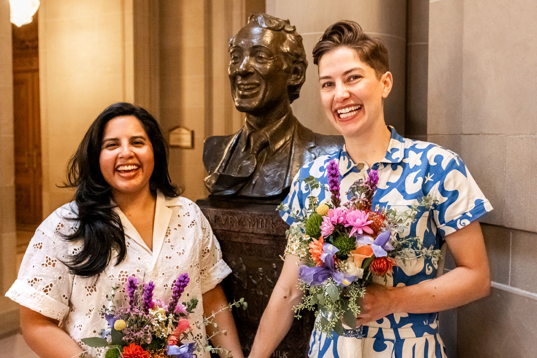 a couple holding hands and laughing standing next to the bust of harvey milk in san francisco city hall