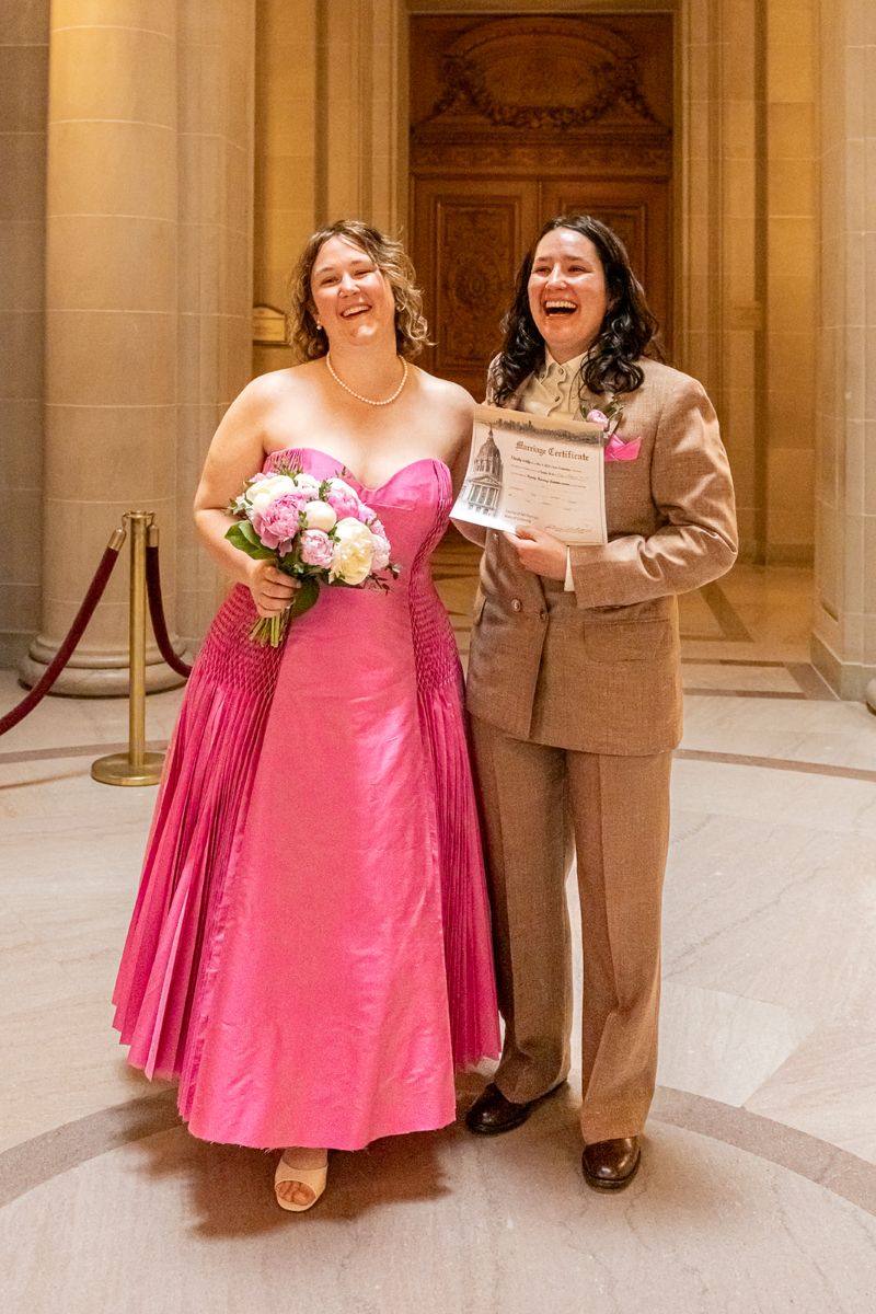 a couple holding their wedding certificate inside of san francisco city hall one person is wearing a pink dress and holding a bouquet and the other is wearing a brown suit