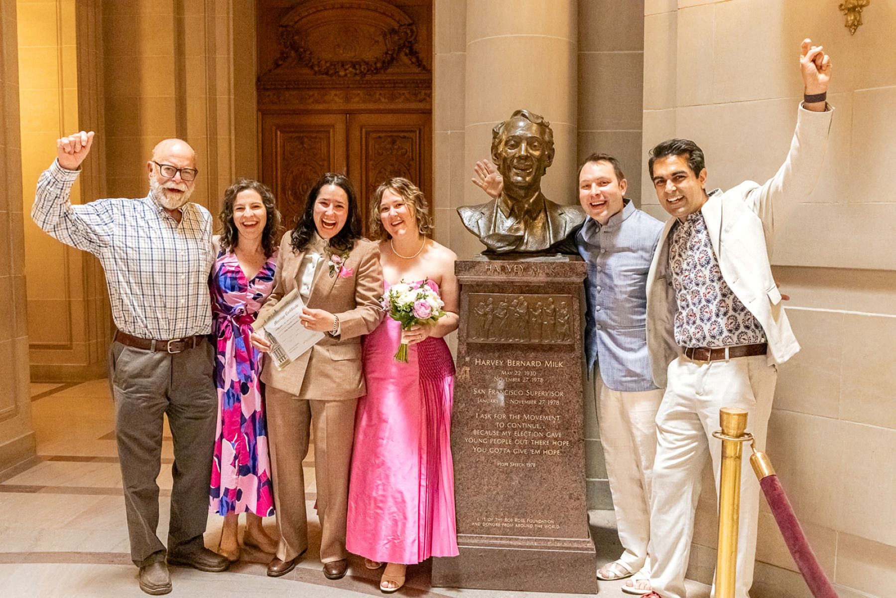a family and a couple taking a picture next to the bust of harvey milk after their wedding ceremony in san francisco city hall