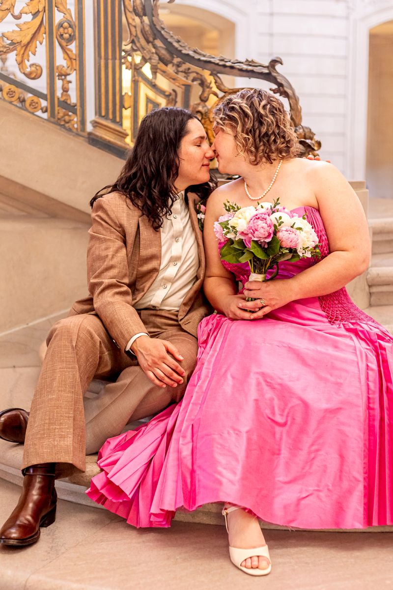 a couple sitting at the bottom of the stairs in san francisco city hall leaning towards each other to kiss one another on the day of their wedding