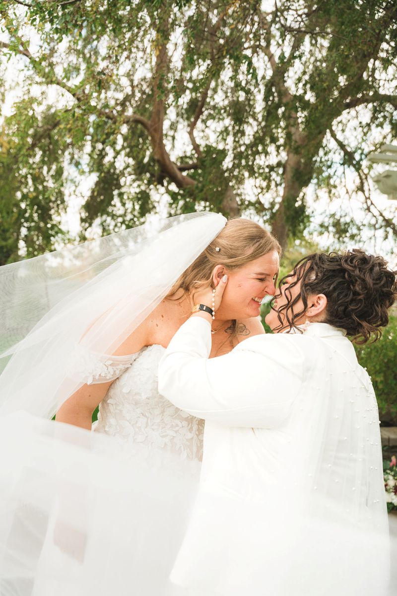 two brides next to a tree leaning in to kiss one another they are both laughing
