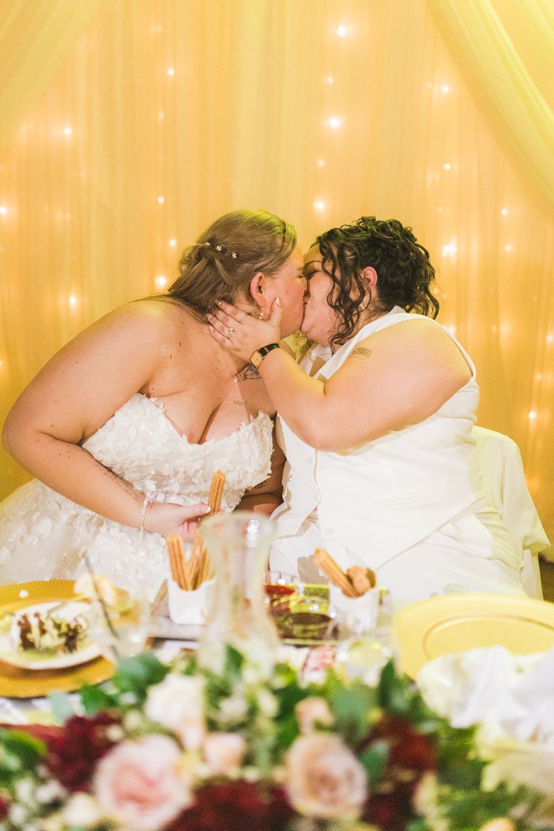 two brides kissing each other at their wedding reception behind them is a curtain with string lights and in front of them are churros