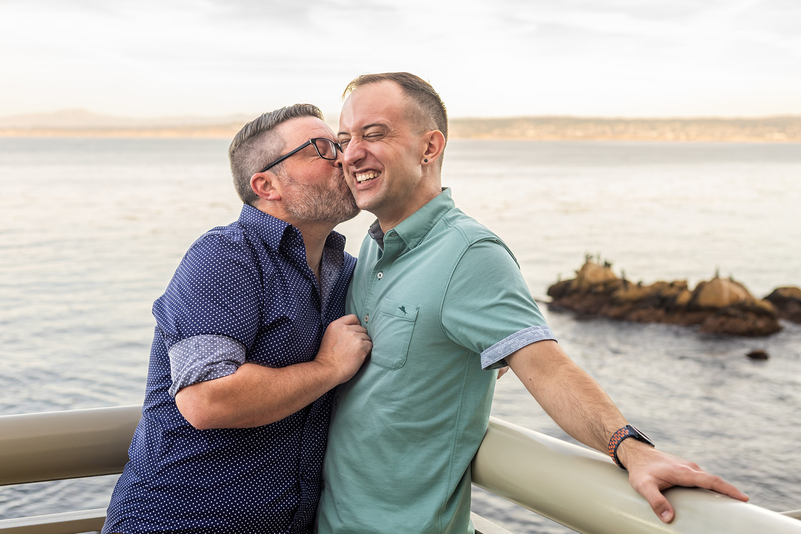 a man giving his partner a kiss on the cheek on the monterey bay aquarium's outer deck next to the ocean